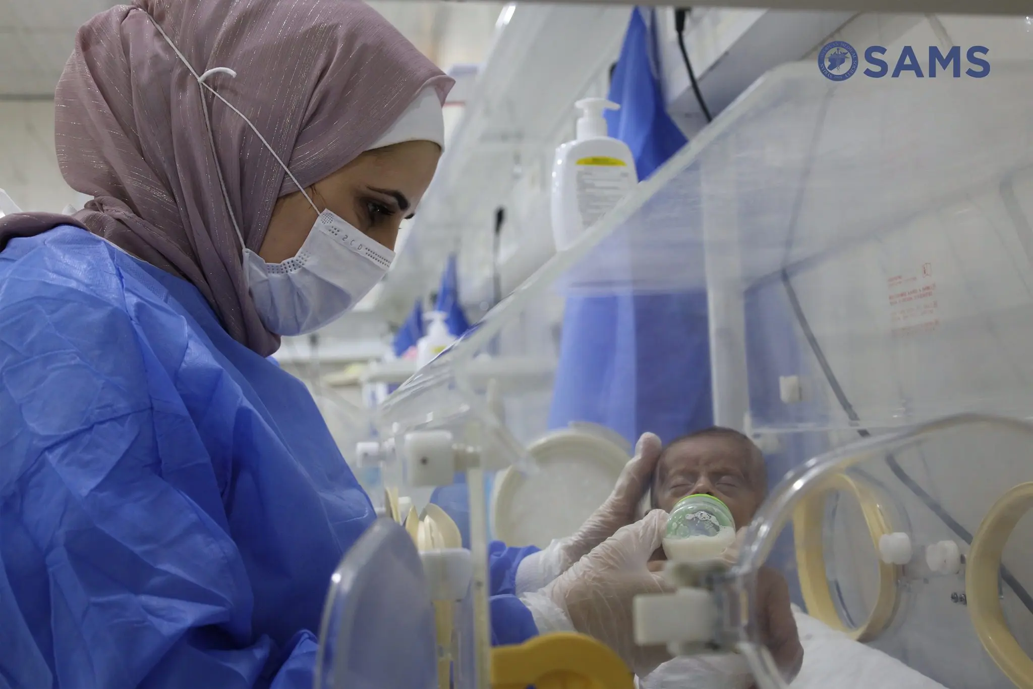 Healthcare worker in a blue protective gown and hijab tending to a newborn in an incubator in a clinical NICU setting