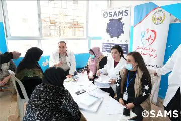 Group of people seated around a table in a bright room, discussing at a SAMS event, with banners in the background and attendees wearing badges and masks.