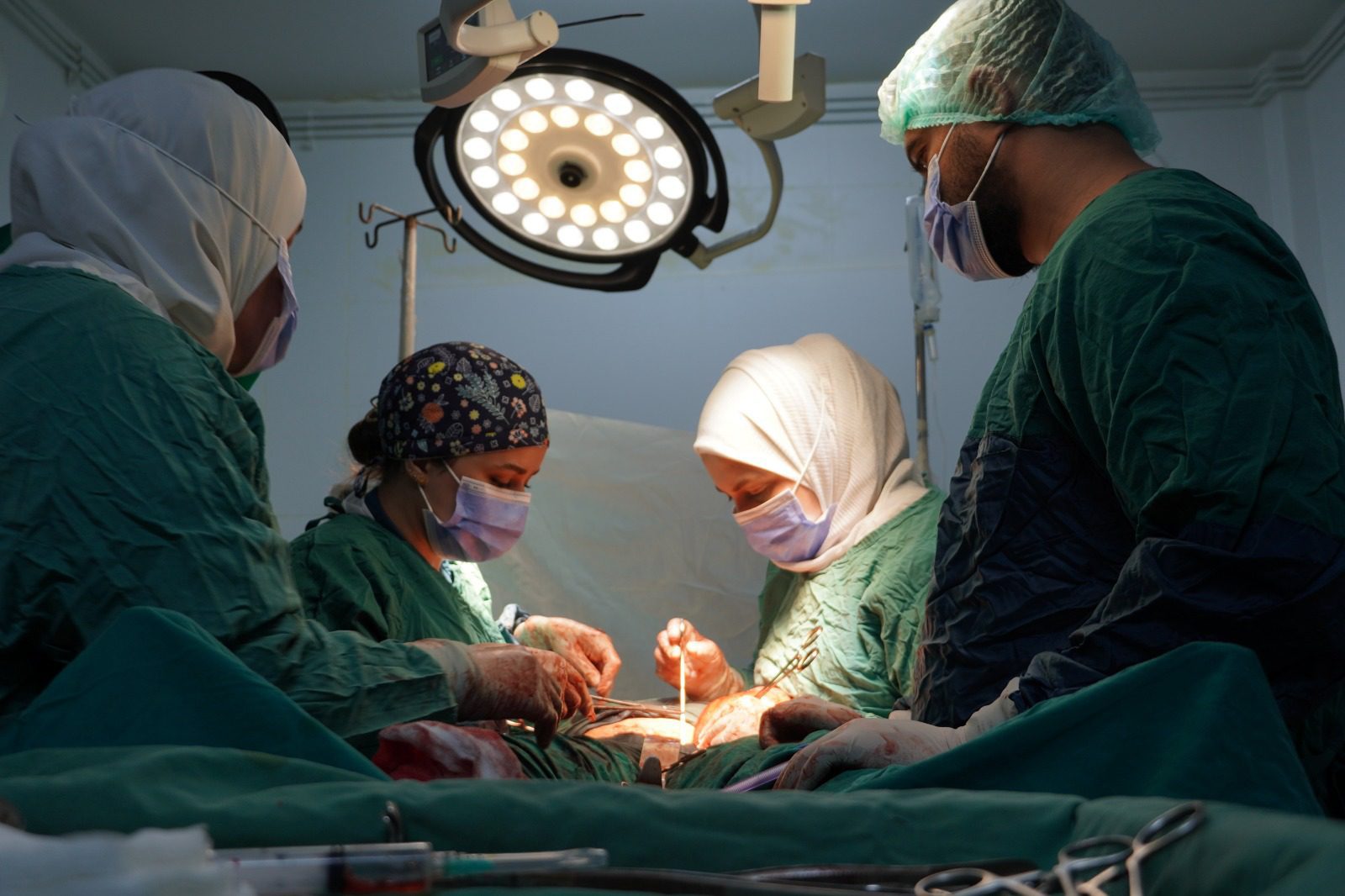 Surgeons in green gowns and masks performing surgery under bright overhead lights in an operating room.